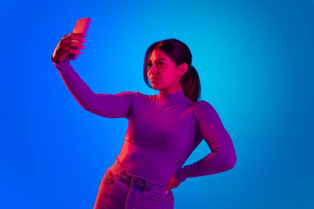 istock Young beautiful girl taking selfie with phone front camera isolated over blue background in pink neon light 1396872864
