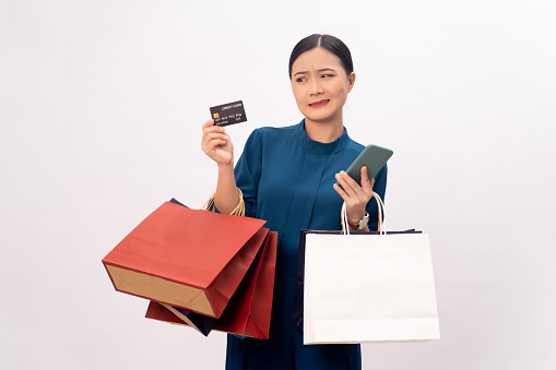 Asian woman guilty worried holding smart phone, credit card and shopping bags mock up standing isolated over white background.