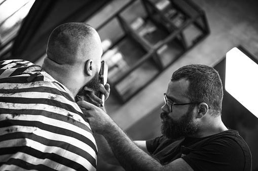 Hipster barber focused on cutting hair in his barber shop. Retro barber shop. Attractive barber cutting hair. Man getting his beard groomed. Black and white photography. Artistic photography.