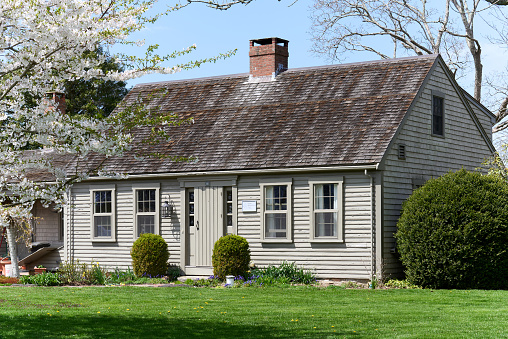 Westport, MA USA - April 23, 2022: A small New England colonial saltbox farmhouse (c.1830) with siding on the front and singles on the side. It is painted a greige color and is shown on a clear day in early spring with a blooming apple tree.