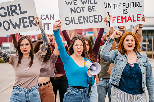 Group of young men and women protesting for equality and women's rights. Holding placards and posters. One young woman talking into the megaphone.