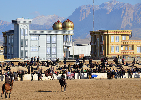 Mazar-e-Sharif, Balkh province, Afghanistan: seated and mounted spectators at a match of Buzkash aka Afghan polo, Afghanistan's national sport - horse-mounted players fight for a calf carcass - the arena is not gated, admission is open and free - mountains in the background and local architecture.