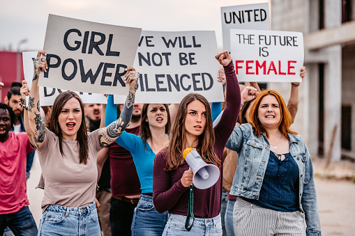 Group of young men and women protesting for equality and women's rights. Holding placards and posters. One young woman talking into the megaphone.