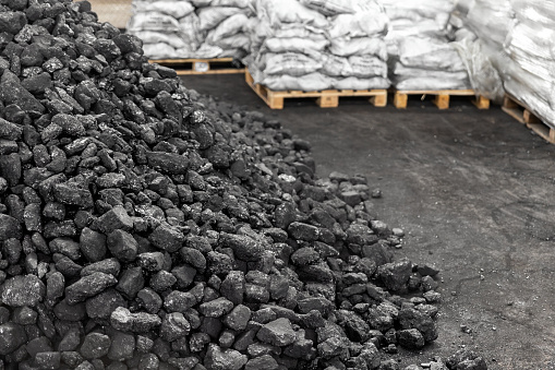 Big heap of dark black lump coal on floor bulk. Charcoal sorage at warehouse stock reserve. activated anthracite packed in plastic bag sack on wooden pallet. Industrial and mining industry background.