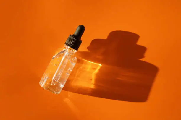 Transparent glass dropper bottle with air bubbles on orange background in sunlight. Pipette with fluid hyaluronic acid, serum, retinol. Cosmetics and healthcare concept Flat lay. Luxury beauty product