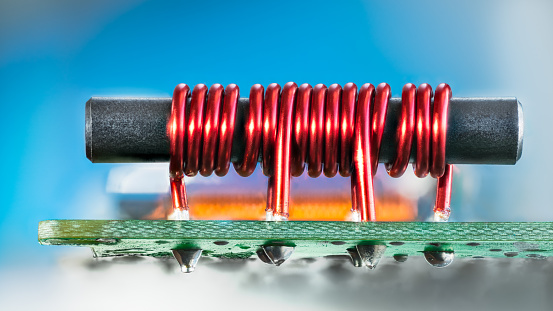 Closeup of red cylindric inductor soldered on green circuit board with tin on bottom side and blue background. Cable TV amp