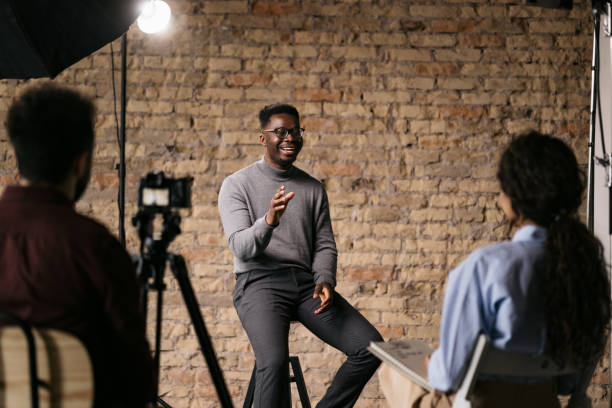 Giving an interview in a modest studio Handsome man sitting on a bar stool and giving an interview to a female journalist in front of him, gesturing with his hands while he's talking and smiling film crew stock pictures, royalty-free photos & images