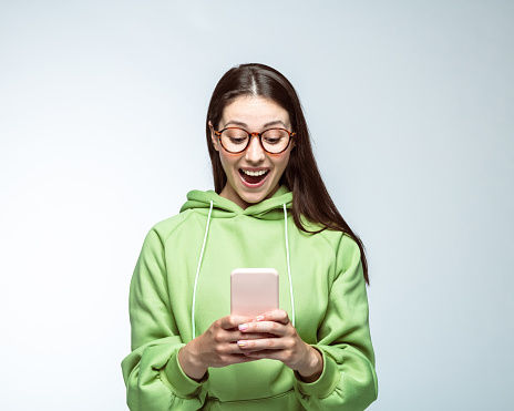 Happy young woman wearing eyeglasses using smart phone with mouth open against white background.