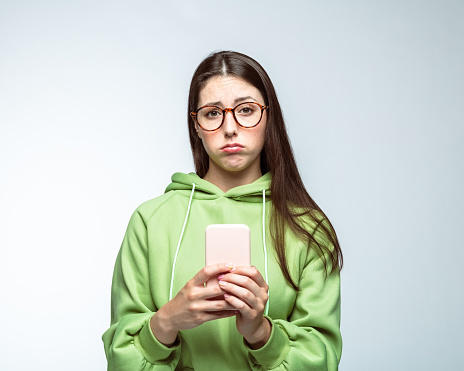 Sad young woman with long brown hair holding mobile phone while standing against white background
