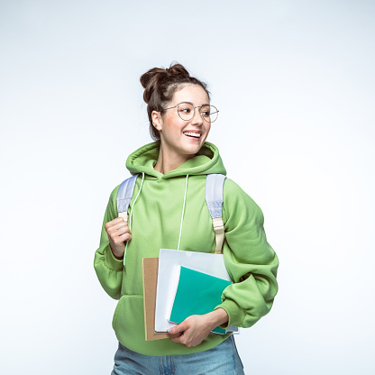 Happy beautiful woman in hair bun and green hoodie standing with backpack and books against white background.