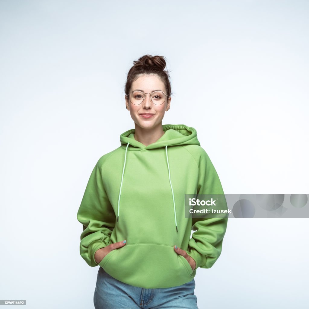 Woman standing with hands in pockets Portrait of smiling confident woman wearing green hooded shirt standing with hands in pockets against white background. Cut Out Stock Photo