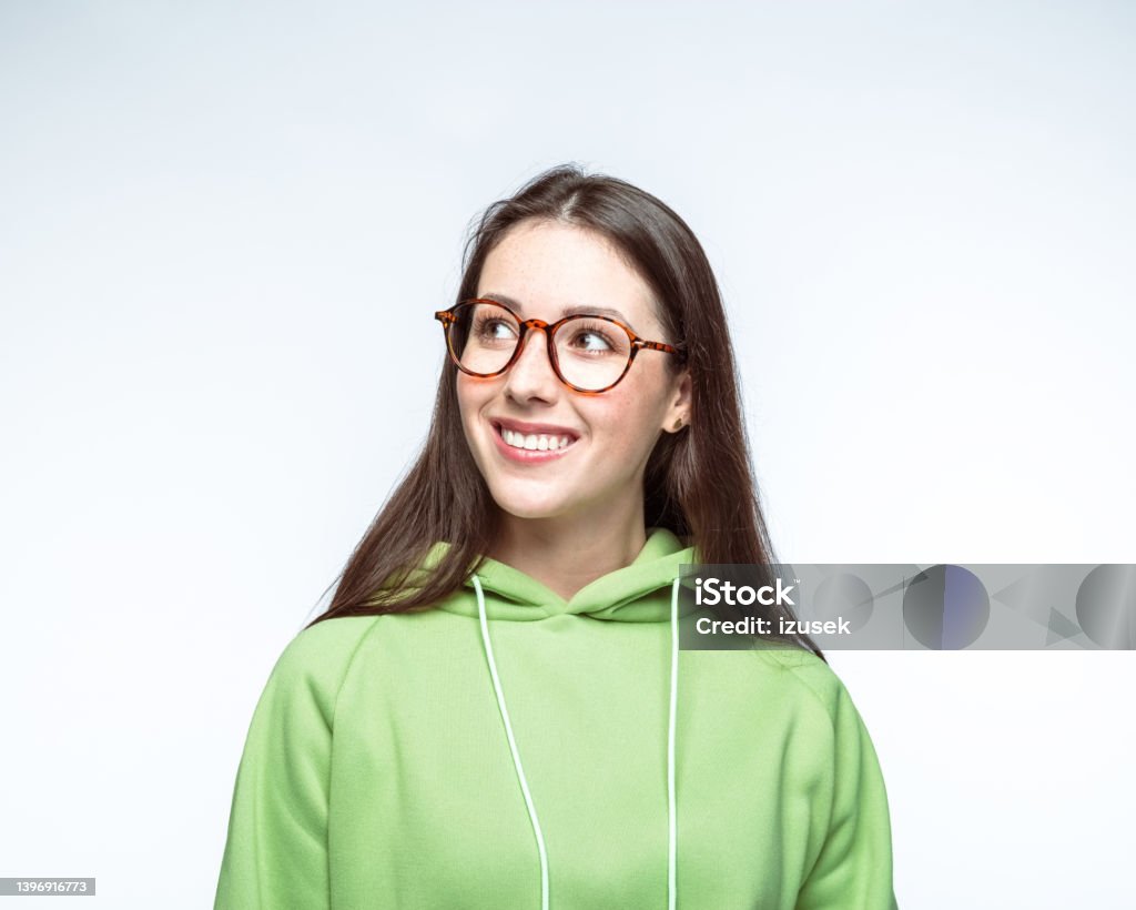Woman wearing eyeglasses looking away Happy young woman wearing eyeglasses and green hoodie looking away while standing against white background. Adult Stock Photo