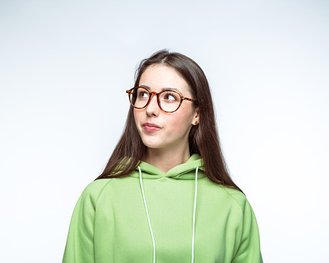 Young beautiful woman with brown hair wearing eyeglasses looking away while standing against white background.