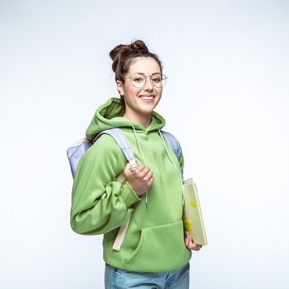 Portrait of happy young woman wearing eyeglasses and green hoodie standing with backpack and books over white background.