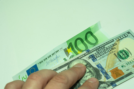 Two banknotes are pressed by the fingers of the hand. 100 dollars and one hundred euros on a light green background.