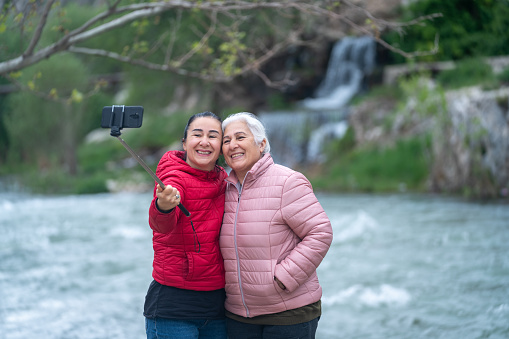 Photo of happy senior mother and daughter taking selfie photo via smartphone attached to monopod. Daughter is wearing a red coat while senior mother is wearing a pink one. Shot in outdoor with a full frame mirrorless camera.