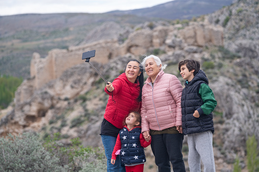 Photo of mother, grandmother, grandson and granddaughter taking selfie photo in outdoor. Mother is wearing a red coat, grandmother is wearing a pink one while little girl and boy is wearing  black coats. Shot in outdoor with afull frame mirrorless camera.