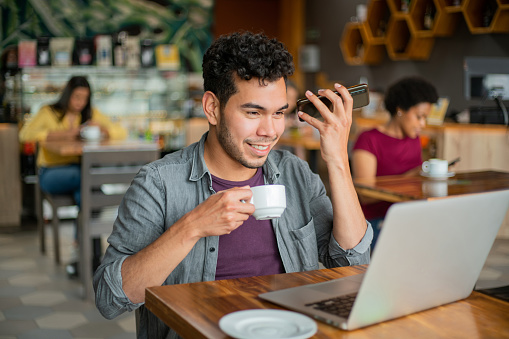 Happy Latin American man at a cafe listening to a voice message on his cell phone while drinking a cup of coffee