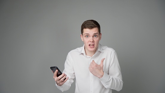 A guy in a white shirt on a gray background, holds a phone in one hand, gestures with his other hand.