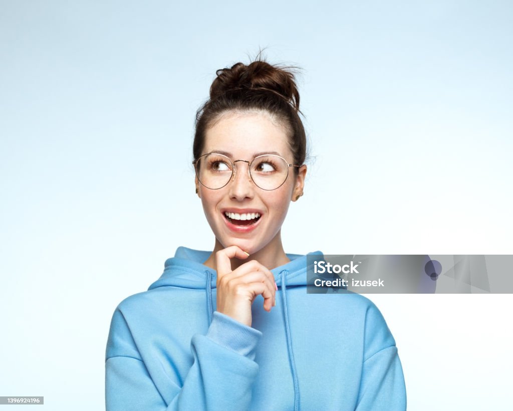 Thoughtful young woman in hooded shirt Smiling young woman wearing blue hoodie with hand on chin against white background. Eyeglasses Stock Photo