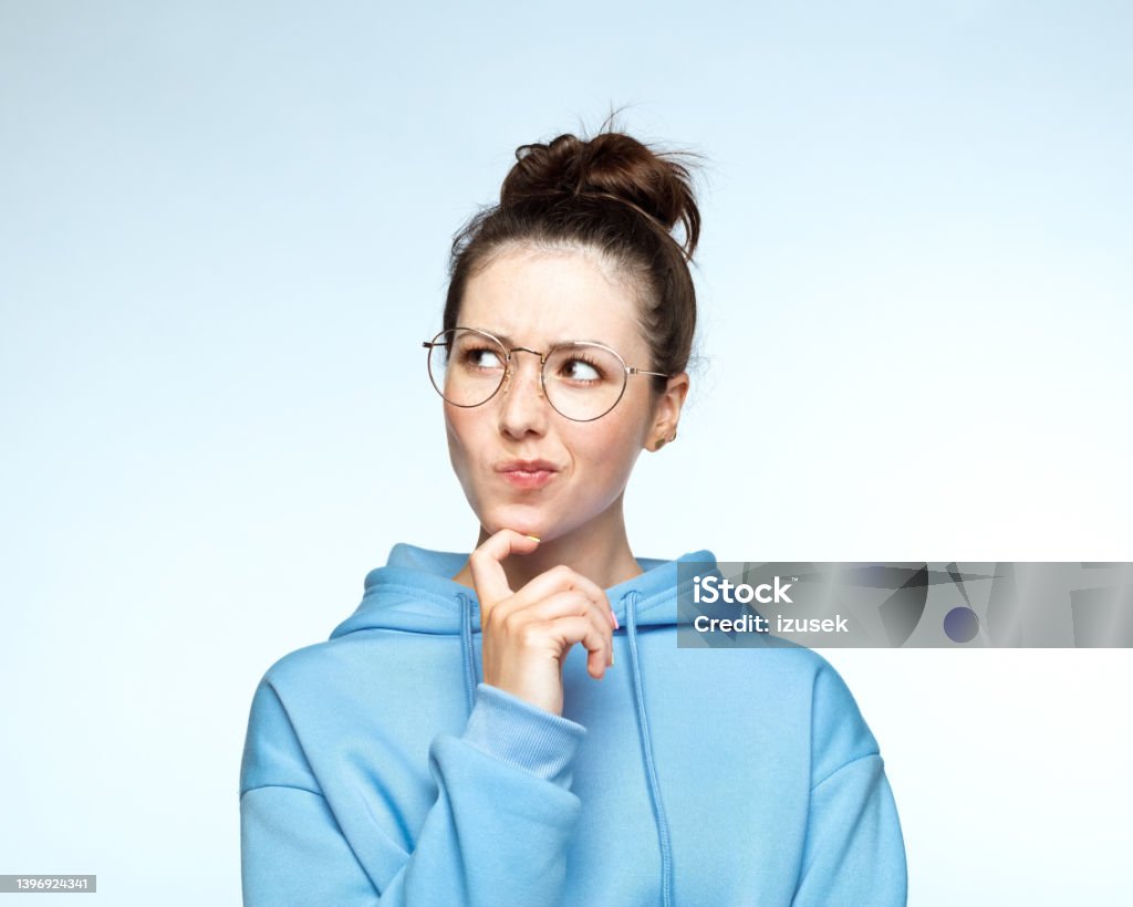 Woman in blue hooded shirt with hand on chin Confused woman with hand on chin puckering lips against white background. 20-24 Years Stock Photo