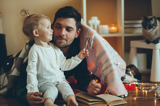 Young father reading book to his little daughter. They built fort made of blankets and decorated it with garland.
