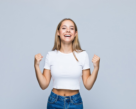 Portrait of happy successful teenage girl in t-shirt clenching fists against white background.