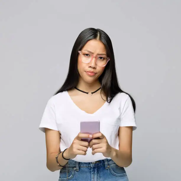 Portrait of confused young woman with mobile phone standing against gray background
