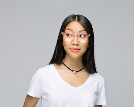 Curious young woman in eyeglasses looking away while standing against gray background