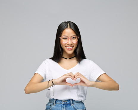 Portrait of happy young woman gesturing heart shape while standing against gray background