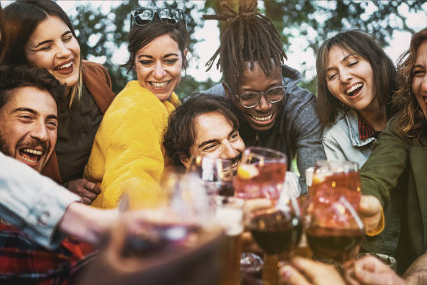 multiethnic group of excited young people having fun toasting wine and beers together at terrace birthday party - lifestyle concept of multigenerational diverse family stay together - focus center man - wine dinner party drinking toast imagens e fotografias de stock