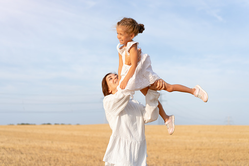 Mother throws up little daughter in air standing wheat field summer day blue sky background. Mom throw up little girl. Happy family concept. White dress. Happy childhood. 35 and 5 years female outdoor