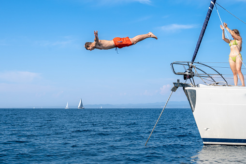 Young man in swimsuit flying with outstretched arms in air above sea surface. Shot in horizontal positing during jumping from yacht. Summer vacation, youth and fun concept.
