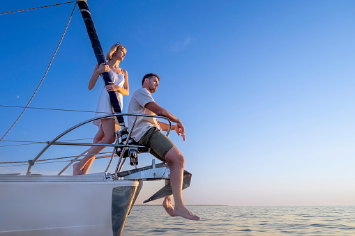 Side view of man sitting on railings and woman standing behind him. Young Caucasian couple on yacht bow in golden hour. Summer vacation, youth and fun concept.