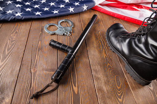 silver metal handcuffs, black ankle boots and police nightstick near crumpled US flag on wooden surface