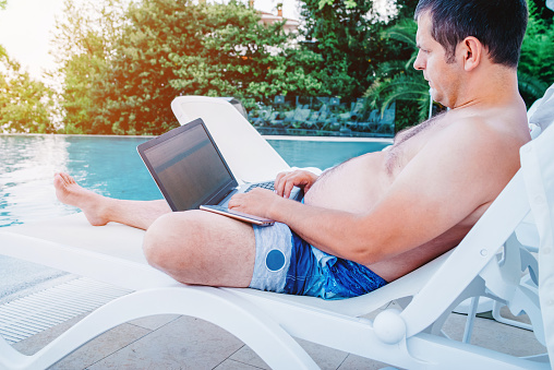 A man on vacation works on a computer by the pool.Rest and work on vacation.Remote work on vacation.Man freelancer working on laptop computer,keyboarding text on beach.Technology and travel.Working outdoors.Freelance concept.