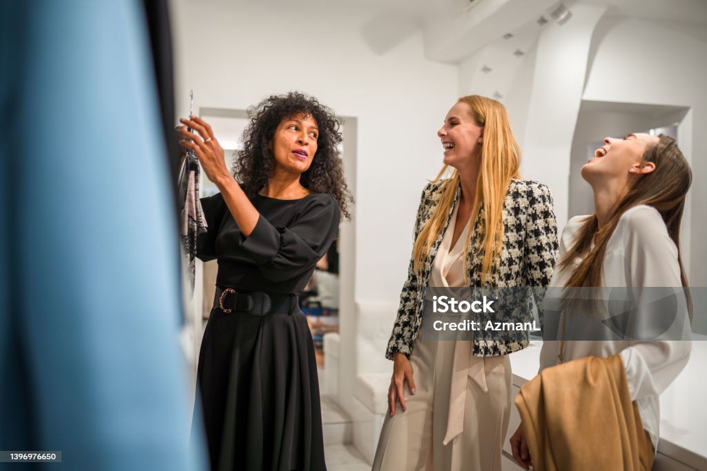 Mid Adult Female Shop Manager With Young Shoppers Two pretty female customers at a clothes shop, the beautiful multiracial owner showing them some garments while they talk and laugh. 3/4 length shot, all looking away. Haute Couture Stock Photo