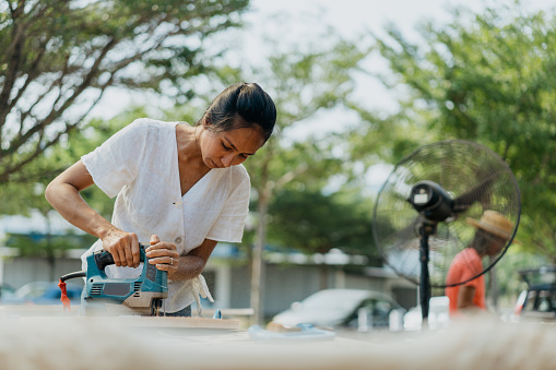 An Asian female carpenter cutting a piece of wood with jigsaw in her workshop