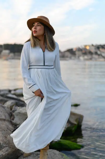 Beautiful woman in long white dress and hat on the rocks by the sea looking into the distance