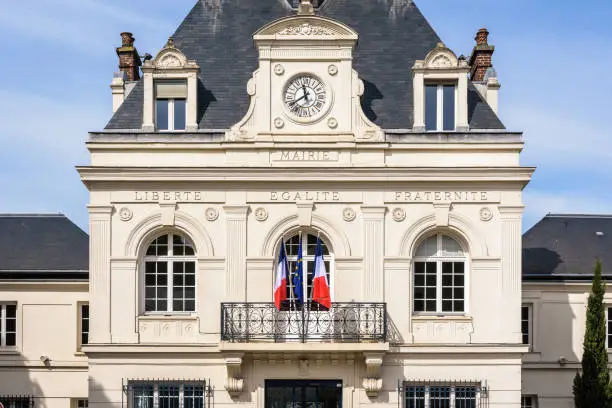 Front view of a french city hall with french and european flags and the national motto of France "Liberty, Equality, Fraternity" engraved on the neoclassical facade under a pediment with clock.