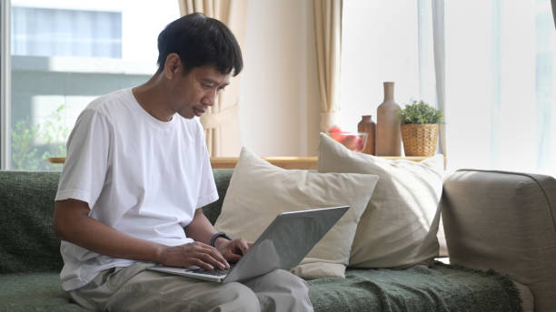 istock Man using laptop, sitting on couch at home, chatting or shopping in internet, reading message, working online with device. 1396997323