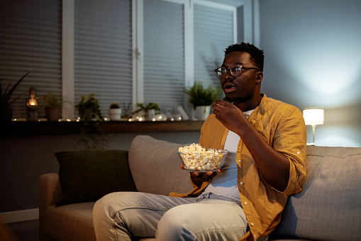 Serious young guy taking some popcorn from a bowl while watching horor movie