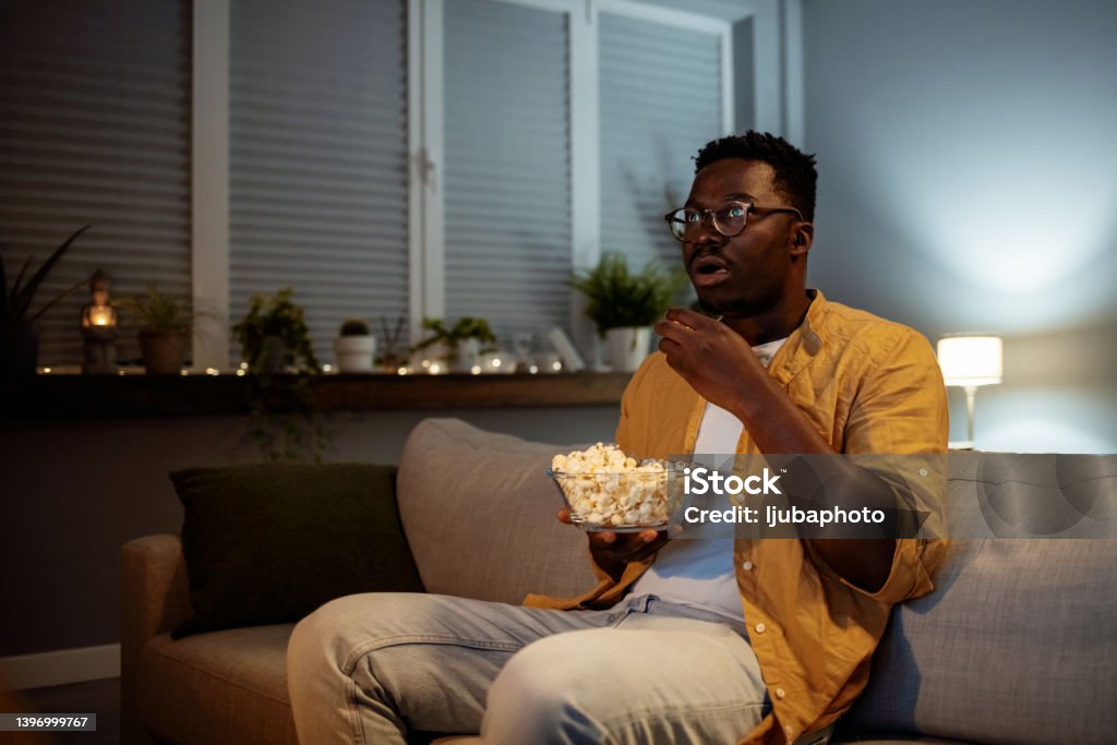 Serious young guy taking some popcorn from a bowl while watching horor movie Television Industry Stock Photo