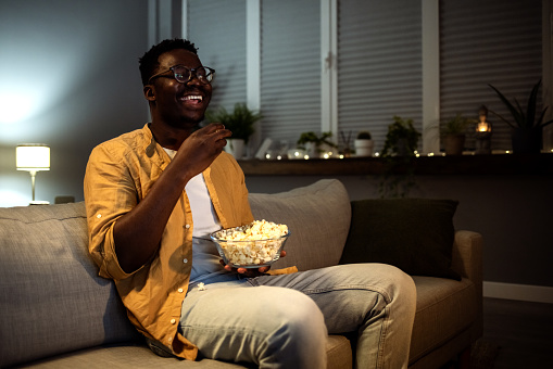 African man sitting at home in his room, watching a movie in the dark, eating popcorn