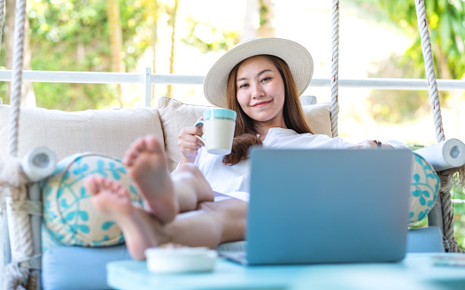 Portrait image of a beautiful young asian woman using laptop computer for remote working or studying online while drinking coffee and laying on swing sofa