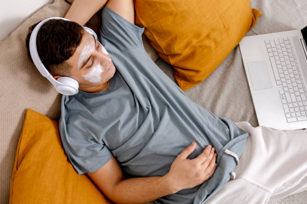 giovane con vestiti casual e maschera di bellezza sul viso sdraiato sul letto con computer portatile e riposo. cosmetico per uomo, cura della pelle. uomo che ascolta musica o audiolibro. rilassarsi a casa. - moisturizing cream audio foto e immagini stock