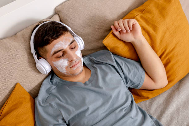 giovane con abiti casual e maschera di bellezza sul viso sdraiato sul letto a casa e a riposo. cosmetico per uomo, cura della pelle. uomo che ascolta musica o audiolibro. rilassante. tempo per te stesso, salute mentale. - moisturizing cream audio foto e immagini stock