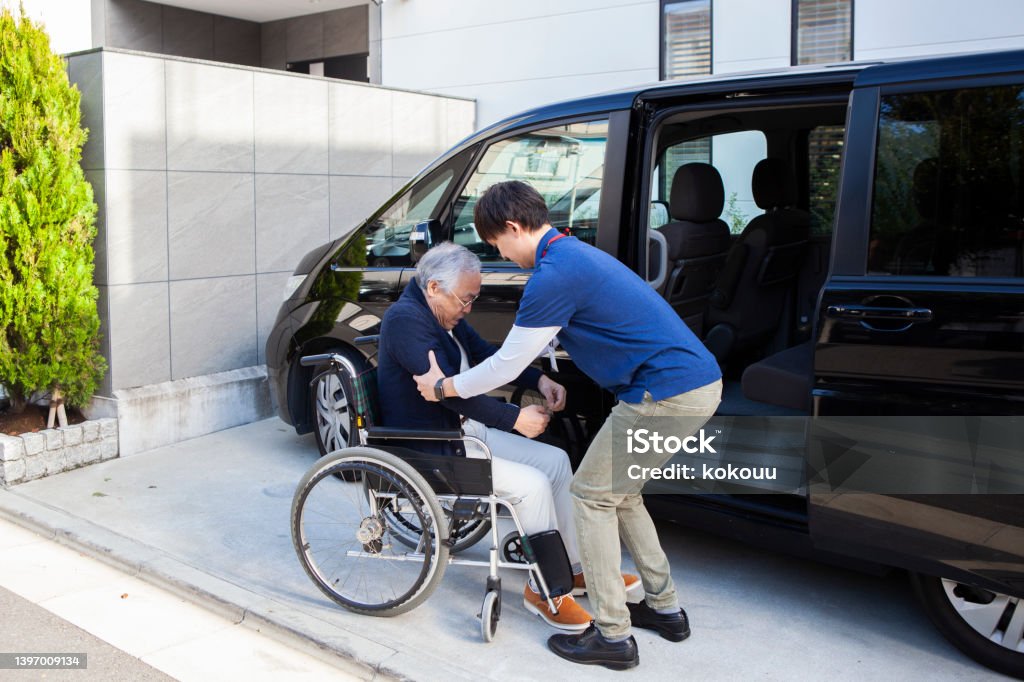 A senior man helped by a young male carer out of car and into wheelchair. An elderly man and public services for active living despite physical disability. Transportation Stock Photo