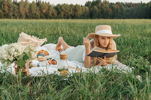 Woman read book on picnic in field. Fruits, cheese, long loaf and tea on a blanket.