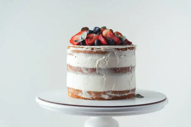Photo of summer vanilla sponge cake with cream cheese filling decorated with strawberries and blueberries on top on a white background. White cake stand
